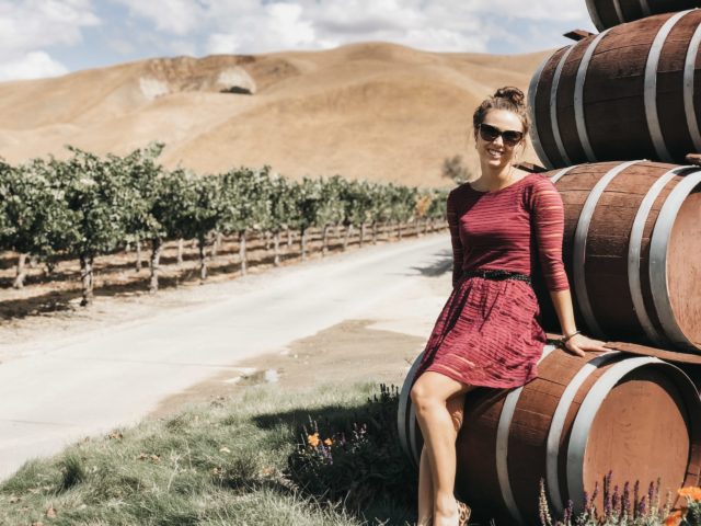a picture of a woman leaning up against a stack of wine barrels with a wine vineyard in the background
